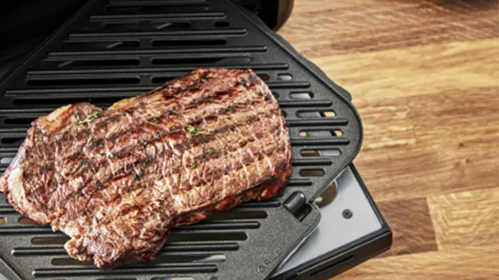 Grillowany stek na czarnym grillu elektrycznym, z drewnianym blatem w tle. Mięso ma widoczne ślady grillowania.