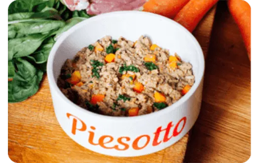 Miska z karmą dla psa "Piesotto" na drewnianej desce. Obok marchewki, szpinak i mięso.