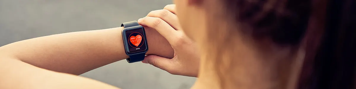 Kobieta sprawdza czarny smartwatch z ikoną czerwonego serca na ekranie. Zegarek na nadgarstku, tło rozmyte.