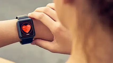 Kobieta sprawdza czarny smartwatch z ikoną czerwonego serca na ekranie. Zegarek na nadgarstku, tło rozmyte.