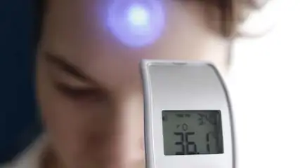 Osoba mierzy temperaturę termometrem bezdotykowym, na ekranie widoczny wynik 36.1 stopni Celsjusza. Tło jest biało-niebieskie.