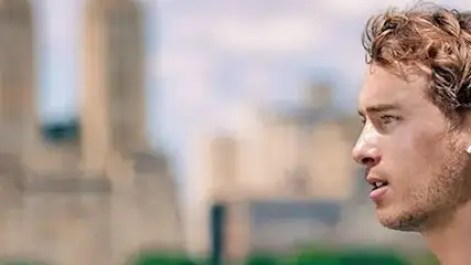 Mężczyzna z słuchawką w uchu, patrzy w lewo, na tle budynków i zieleni.