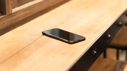 Na drewnianym blacie leży czarny smartfon. W tle widać krzesła barowe i fragment okna.