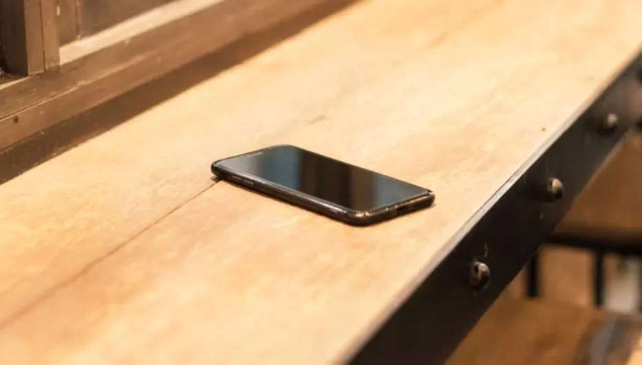 Na drewnianym blacie leży czarny smartfon. W tle widać krzesła barowe i fragment okna.