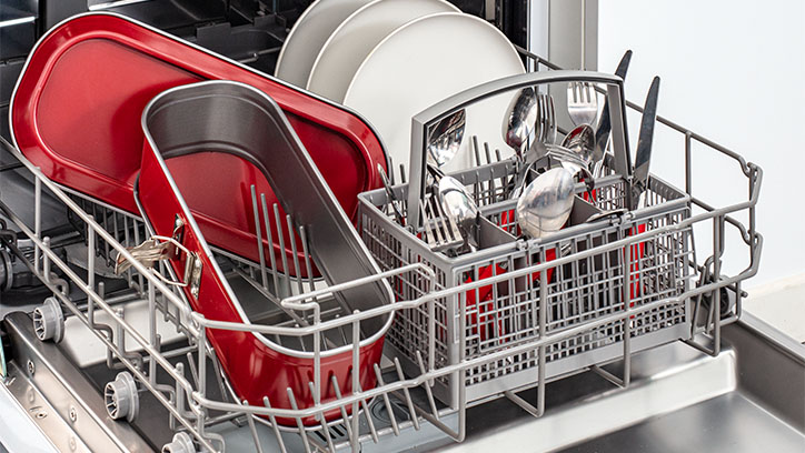 Wnętrze zmywarki z metalowym koszem. W koszu czerwone naczynia, białe talerze, sztućce i metalowe przybory kuchenne.