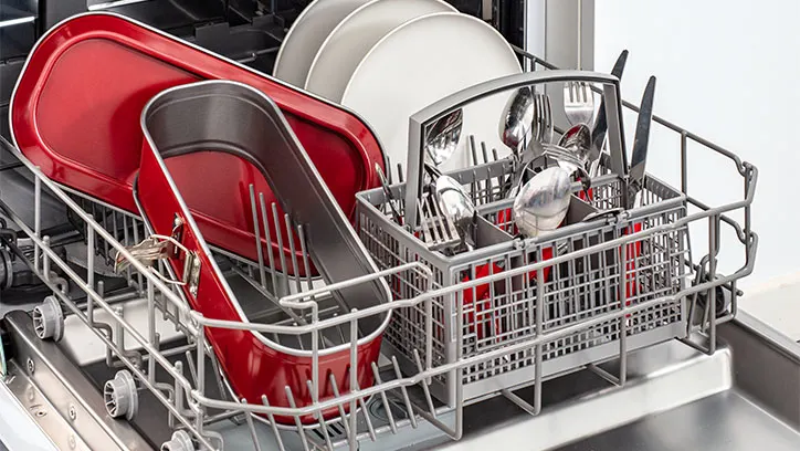 Wnętrze zmywarki z metalowym koszem. W koszu czerwone naczynia, białe talerze, sztućce i metalowe przybory kuchenne.