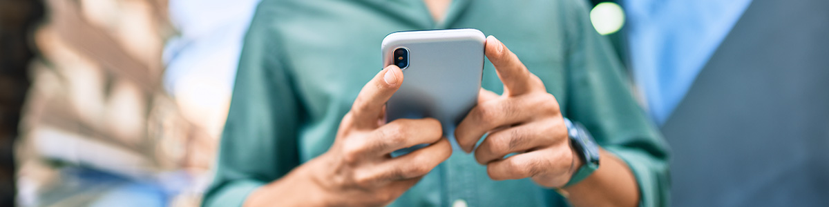 Osoba w zielonej koszuli trzyma szary smartfon obiema rękami. Na nadgarstku widoczny zegarek. Tło jest rozmyte.