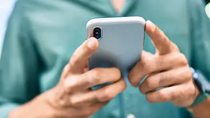 Osoba w zielonej koszuli trzyma szary smartfon obiema rękami. Na nadgarstku widoczny zegarek. Tło jest rozmyte.