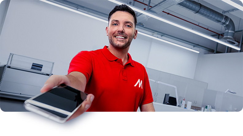 Mężczyzna w czerwonej koszulce polo z logo podaje smartfon. W tle biuro z oświetleniem sufitowym i sprzętem.