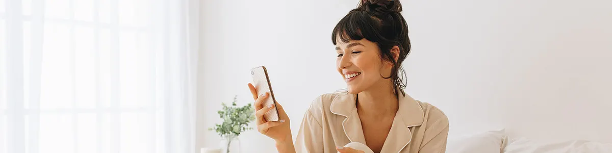 Uśmiechnięta kobieta w piżamie trzyma telefon. W tle wazon z zielonymi roślinami i białe zasłony.