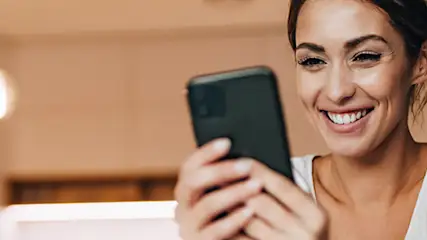 Uśmiechnięta kobieta w białej bluzce trzyma czarny smartfon na tle kuchni z lampami i sprzętem.