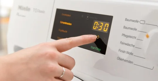 Na białej pralce palec naciska przycisk Start/Stop obok czarnego ekranu z czasem 0:30. Obok widoczne opcje prania.