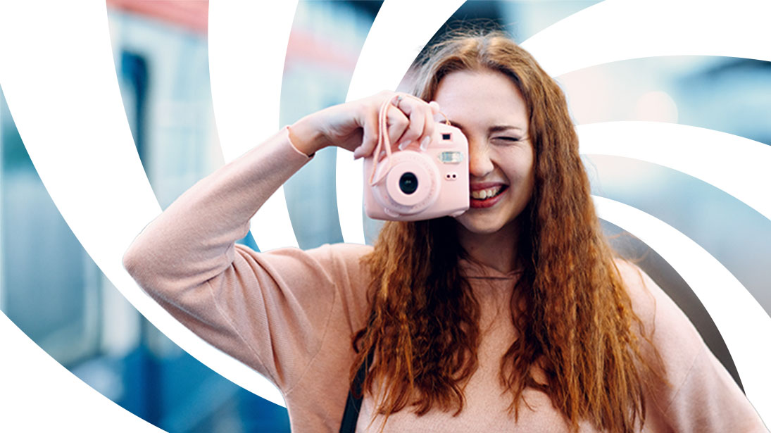 Uśmiechnięta, młoda kobieta z rudymi włosami trzyma różowy aparat instax i celuje obiektywem w stronę widza.