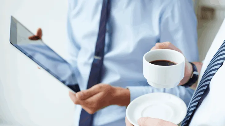 Mężczyzna trzymający laptopa, obok drugi mężczyzna trzymający kawę, oboje mają krawaty i koszule.