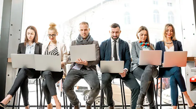 Pięć osób siedzi na krzesłach i patrzy w laptopy; w tle okno.