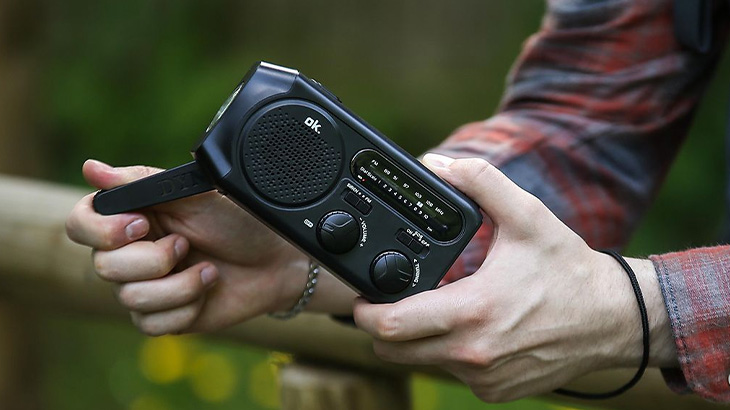 Osoba trzyma czarne radio z anteną na tle zieleni. Radio ma głośnik, pokrętła i skalę częstotliwości.