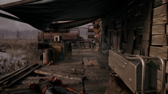 Na zdjęciu widoczny jest zrujnowany drewniany budynek z beczkami na zewnątrz i widokiem na zarośnięty teren. Widać również wózek.