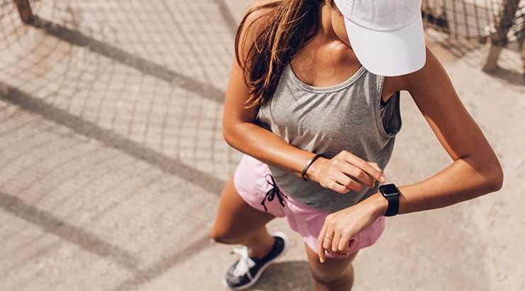 Kobieta w szarej koszulce i różowych szortach sprawdza zegarek na chodniku. W tle siatka i cień.