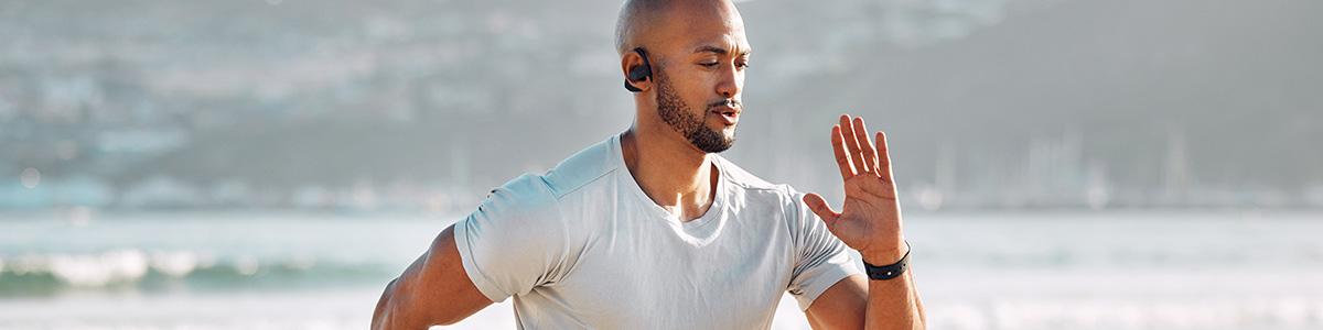 Mężczyzna z słuchawkami biegnie na tle plaży i morza, unosząc dłoń. Nosi białą koszulkę i czarny zegarek.