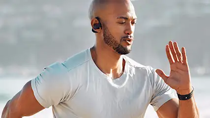 Mężczyzna z słuchawkami biegnie na tle plaży i morza, unosząc dłoń. Nosi białą koszulkę i czarny zegarek.