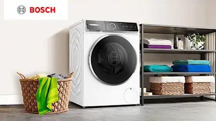 Widok na pralkę obok drewnianej szafki w kuchni, z szarymi szafkami z metalowymi uchwytami w tle.