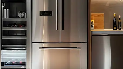 Wnętrze kuchni ze stalową lodówką i szafkami. Na zewnątrz widoczny ogród z zielenią i szarą ławką.