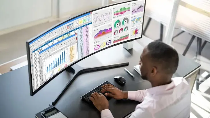 Osoba pracuje przy biurku z monitorem panoramicznym, na którym widoczne są wykresy i dane. Używa klawiatury i myszy.