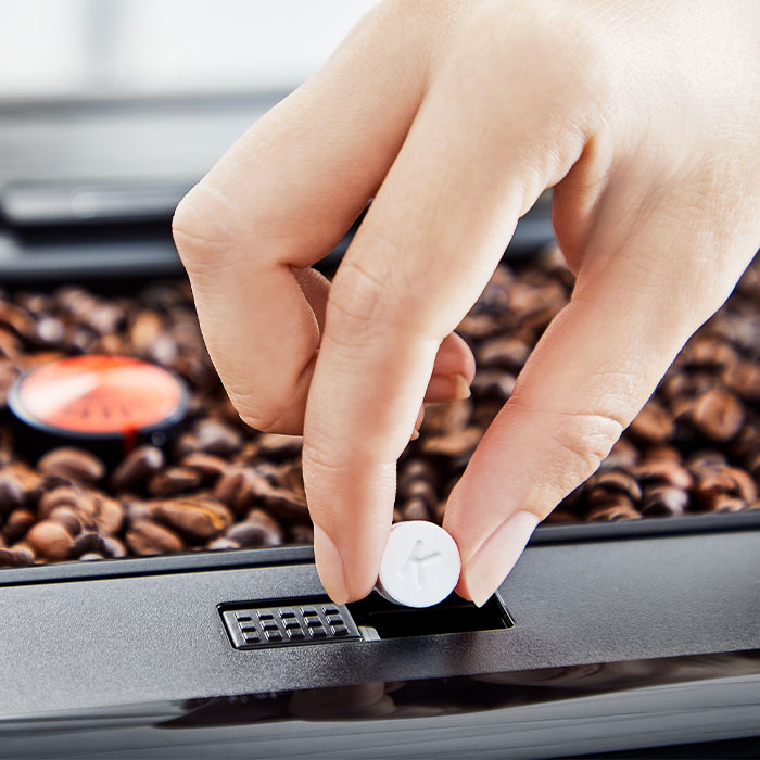 Dłoń umieszcza białą tabletkę w czarnym urządzeniu. W tle widoczne ziarna kawy i pomarańczowa kapsułka.