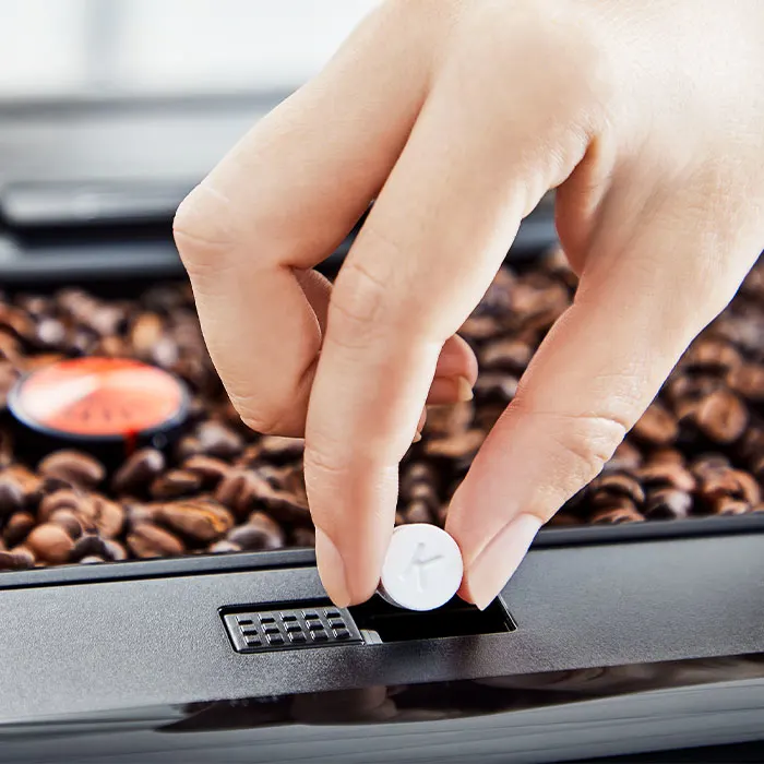 Dłoń umieszcza białą tabletkę w czarnym urządzeniu. W tle widoczne ziarna kawy i pomarańczowa kapsułka.