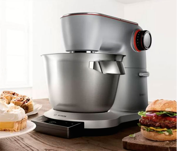 Srebrny robot kuchenny Bosch z misą. Na stole ciasto i burger.