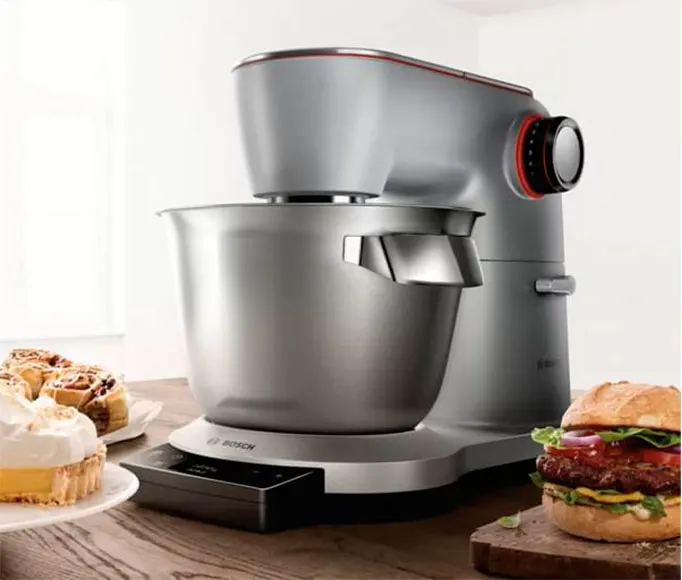 Srebrny robot kuchenny Bosch z misą. Na stole ciasto i burger.