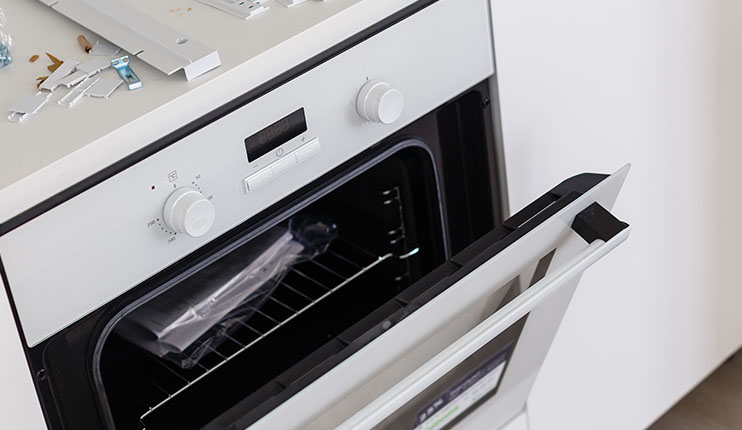 Otwarty, biały piekarnik z rusztem w środku. Na górze piekarnika leżą metalowe elementy montażowe.