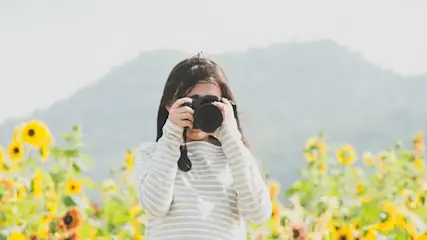 Kobieta robi zdjęcie aparatem na tle pola słoneczników i gór w oddali.