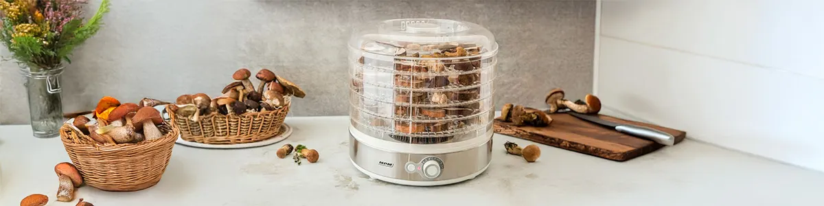Pełna suszarka do grzybów typu modułowego na białym blacie kuchennym, obok grzyby w małych koszyczkach i na desce do krojenia. 
