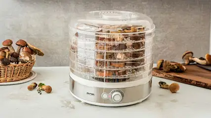 Pełna suszarka do grzybów typu modułowego na białym blacie kuchennym, obok grzyby w małych koszyczkach i na desce do krojenia.