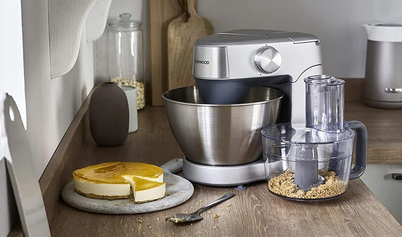 Na drewnianym blacie stoją: robot kuchenny Kenwood, tort serowy, szklany pojemnik i deski do krojenia.