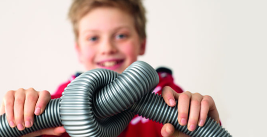 Chłopiec trzyma w dłoniach szary, karbowany wąż, zgięty w kształt węzła na tle białej ściany.