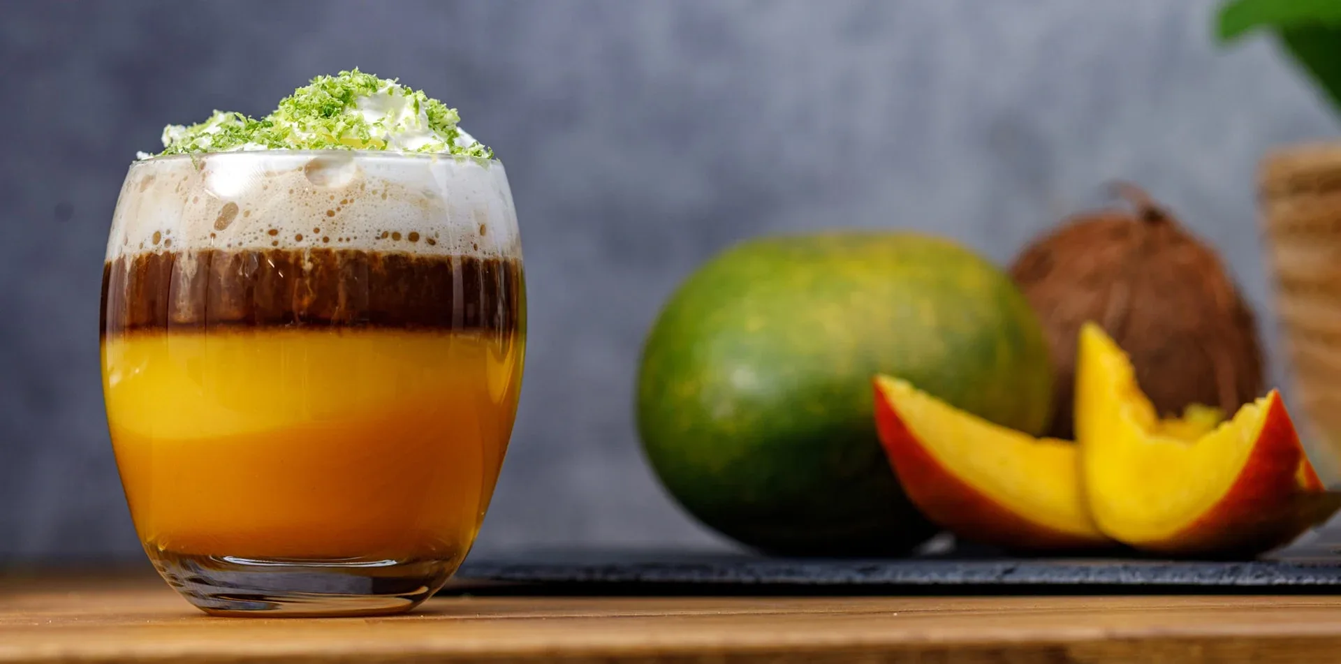 Szklanka z warstwami napoju, mango i kokos na drewnianym stole. Napój ma warstwy pomarańczowe, brązowe i białą piankę z zieloną posypką.
