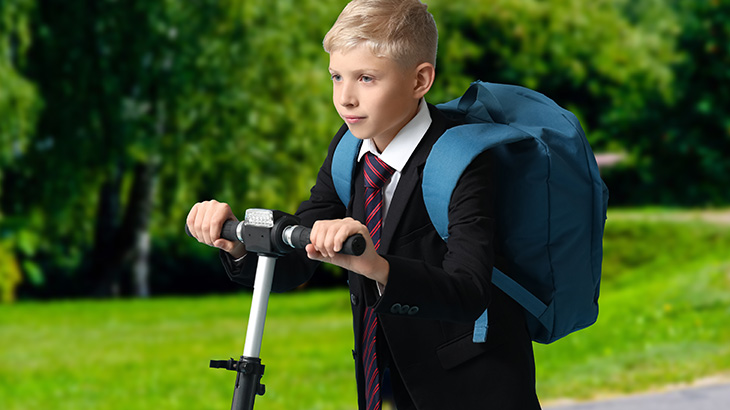 Chłopiec w garniturze i z niebieskim plecakiem jedzie hulajnogą po ścieżce w parku. W tle widać zielone drzewa i trawnik.