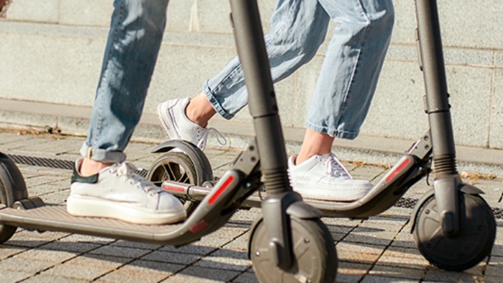 Dwie osoby na hulajnogach elektrycznych. Osoby ubrane w jeansy i białe buty stoją na szarych hulajnogach na chodniku.