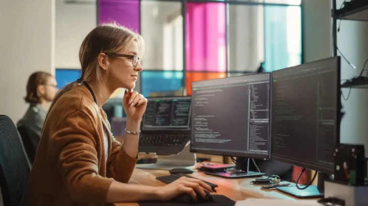 Kobieta w okularach pracuje przy dwóch monitorach z kodem. W tle okno z kolorowymi szybami i inna osoba.