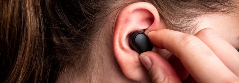 Osoba wkłada czarną słuchawkę bezprzewodową do ucha. W tle włosy.