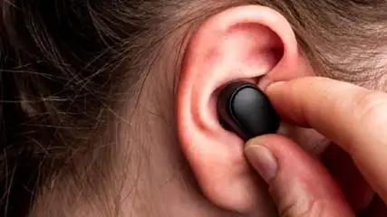 Osoba wkłada czarną słuchawkę bezprzewodową do ucha. W tle włosy.