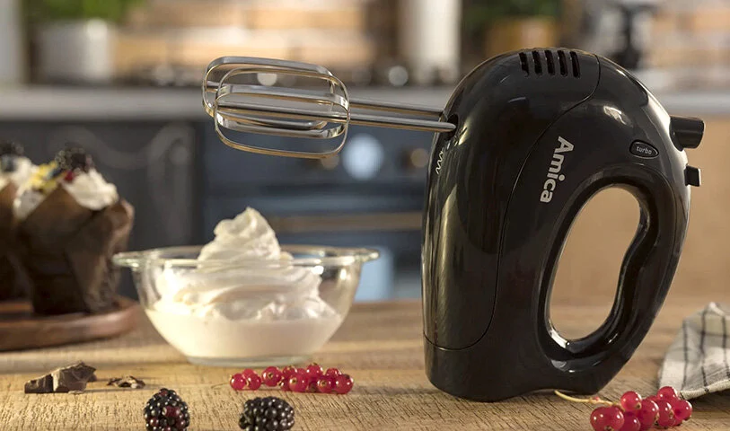 Na drewnianym stole stoi czarny mikser Amica, miska z bitą śmietaną, babeczki, jagody i porzeczki. Tło jest rozmyte.