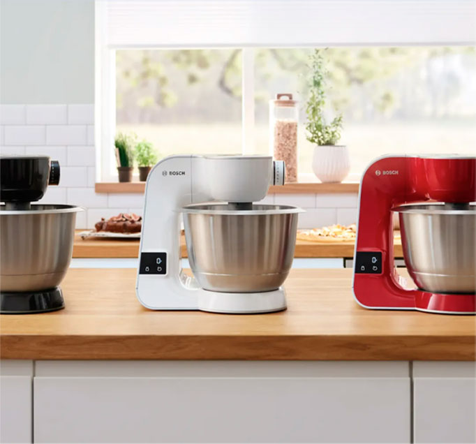 Na blacie stoją trzy miksery Bosch w kolorach: czarnym, białym i czerwonym, z metalowymi misami. W tle okno.