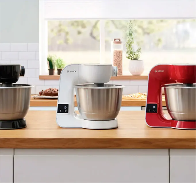 Na blacie stoją trzy miksery Bosch w kolorach: czarnym, białym i czerwonym, z metalowymi misami. W tle okno.