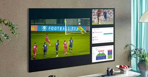 Telewizor pokazuje mecz piłki nożnej i dane. Obok zasłona, roślina i owoce na stole.