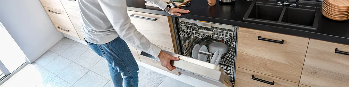 Otwarta zmywarka z srebrnym koszem na sztućce i białymi talerzami wewnątrz, na tle białych szafek kuchennych.