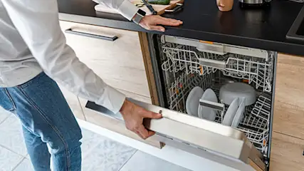 Otwarta zmywarka z srebrnym koszem na sztućce i białymi talerzami wewnątrz, na tle białych szafek kuchennych.