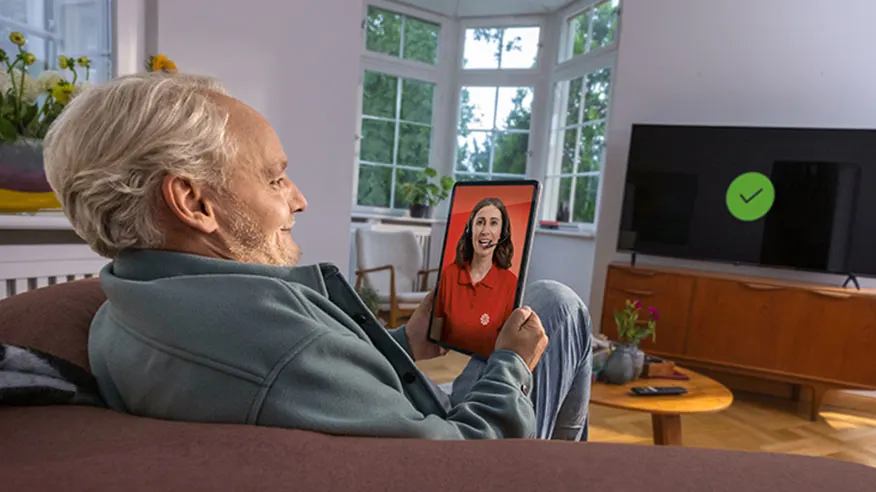 Mężczyzna siedzi na kanapie z tabletem. Na ekranie tabletu kobieta w czerwonej koszuli. Za nim okno i telewizor z zielonym symbolem.